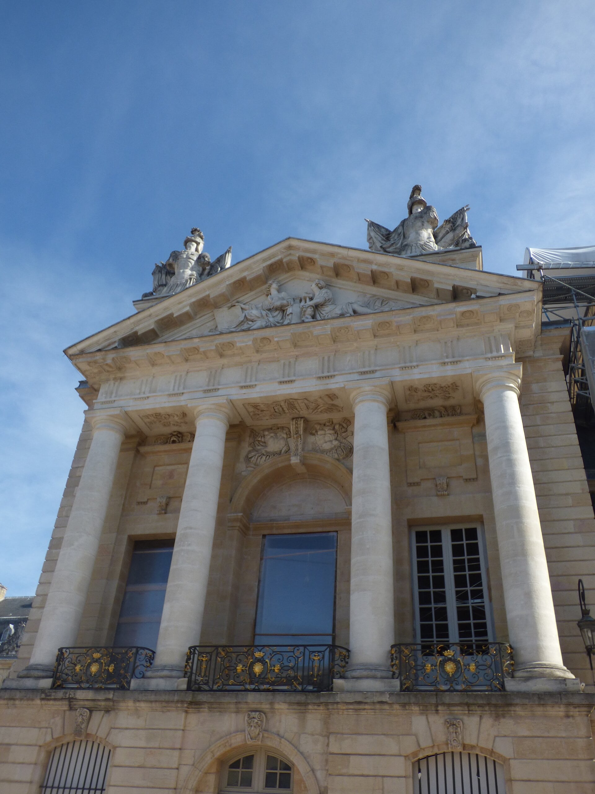 Vue en contre-plongée d’un avant-corps classique : colonnes corinthiennes géantes, fronton triangulaire sculpté de figures allongées, statues d’angle au faîte, balcons de fer forgé rehaussés d’or portant une couronne ducale