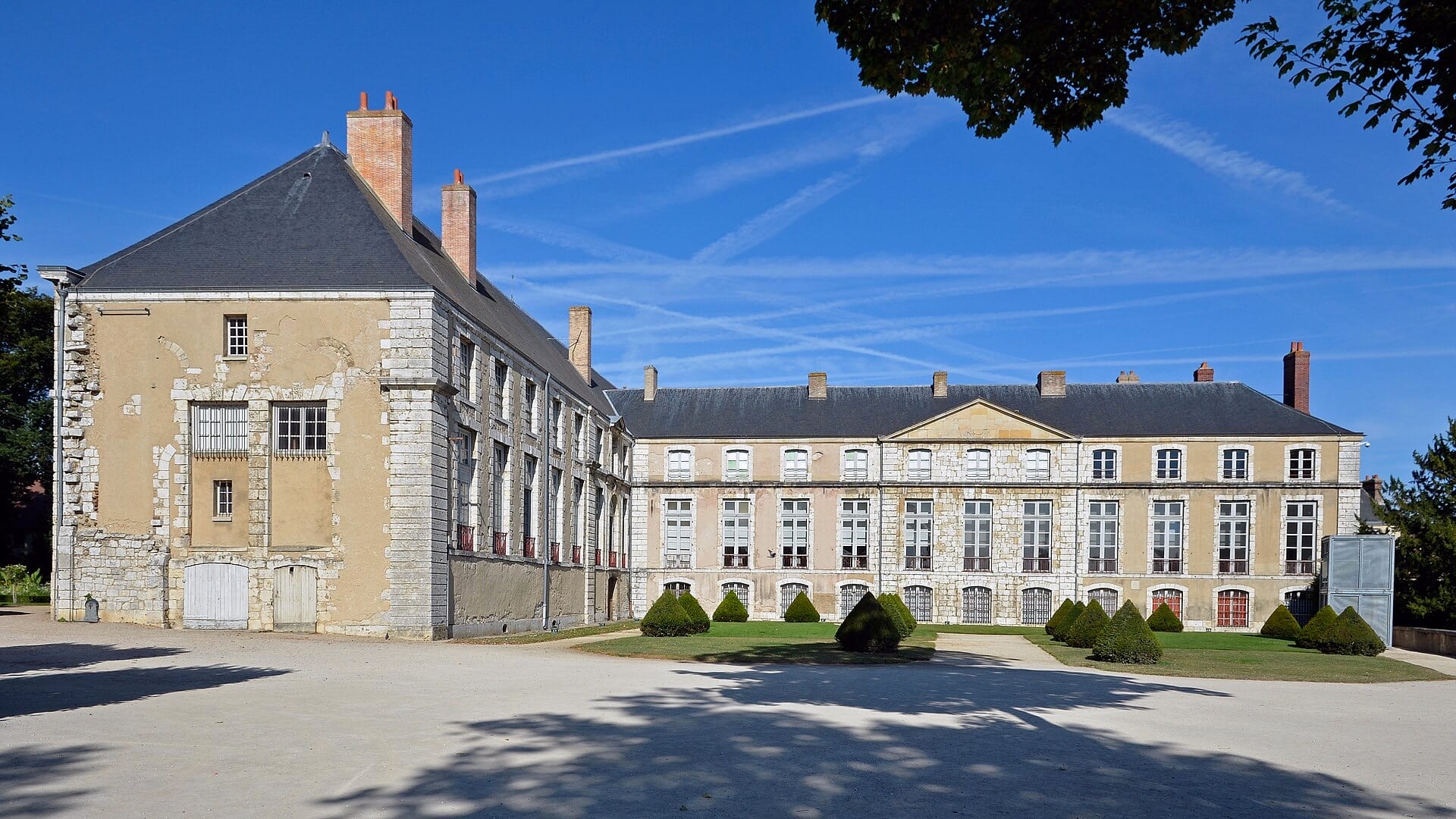 Deux corps de logis anciens en pierre autour d’une cour de gravier : à gauche un pavillon à haut toit d’ardoise et hautes cheminées de brique, à droite une longue aile classique à fronton central ; parterres et ifs taillés