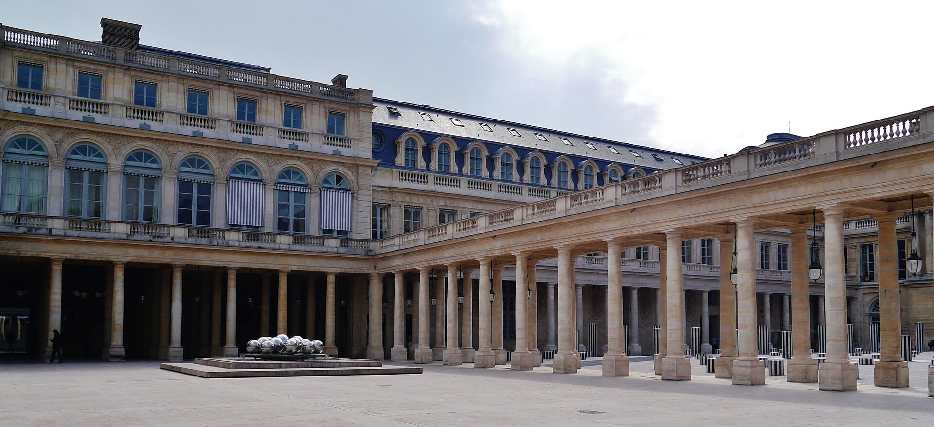 Cour d’un palais classique bordée d’une longue galerie à colonnes toscanes ; à gauche, une aile à grandes fenêtres cintrées ; au sol, une fontaine de sphères métalliques polies