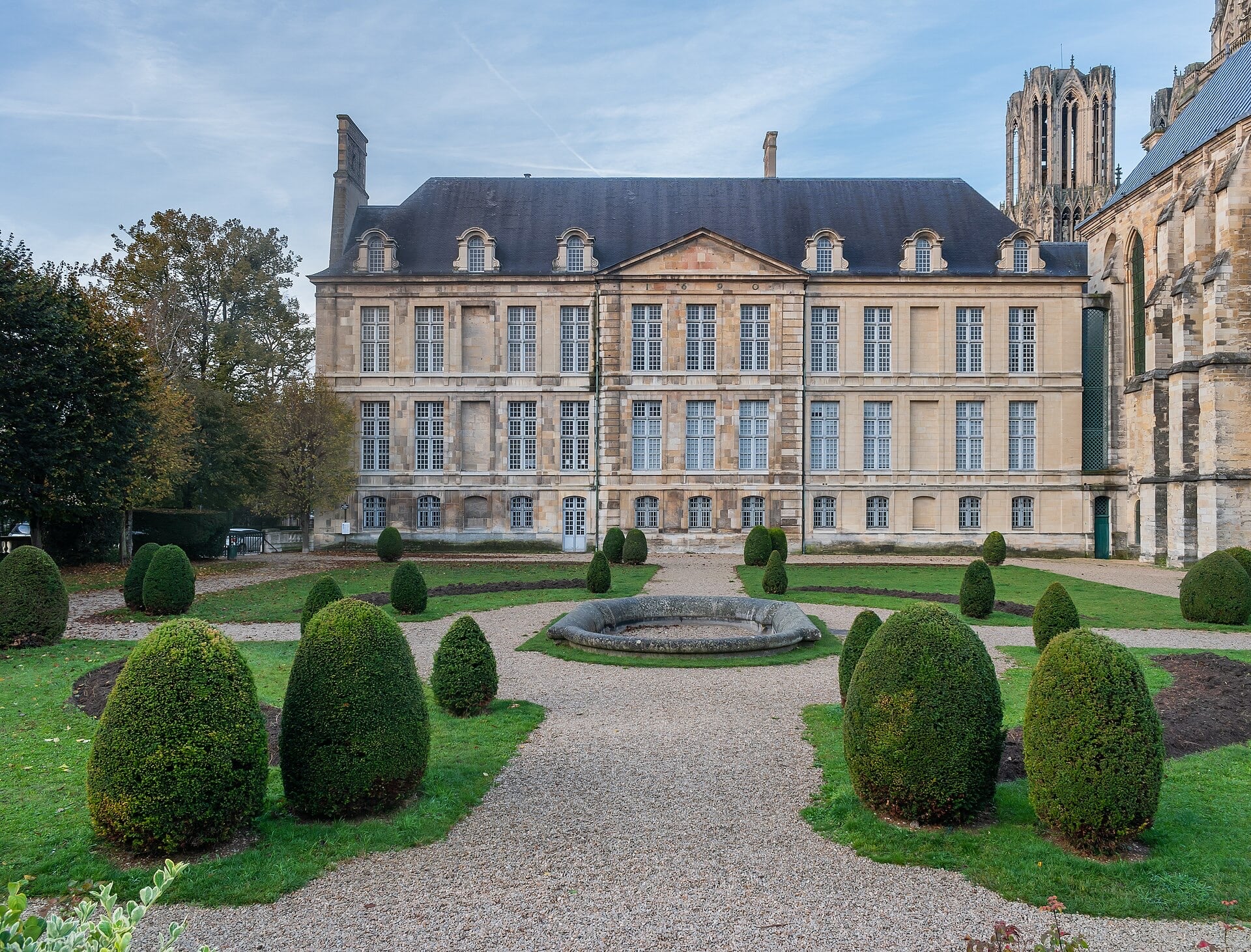 Façade classique en pierre de taille d’un palais à haute toiture d’ardoise et lucarnes à fronton, datée 1690 au fronton central ; devant, un jardin régulier à ifs taillés et vasque de pierre ; à droite, une tour de cathédrale gothique