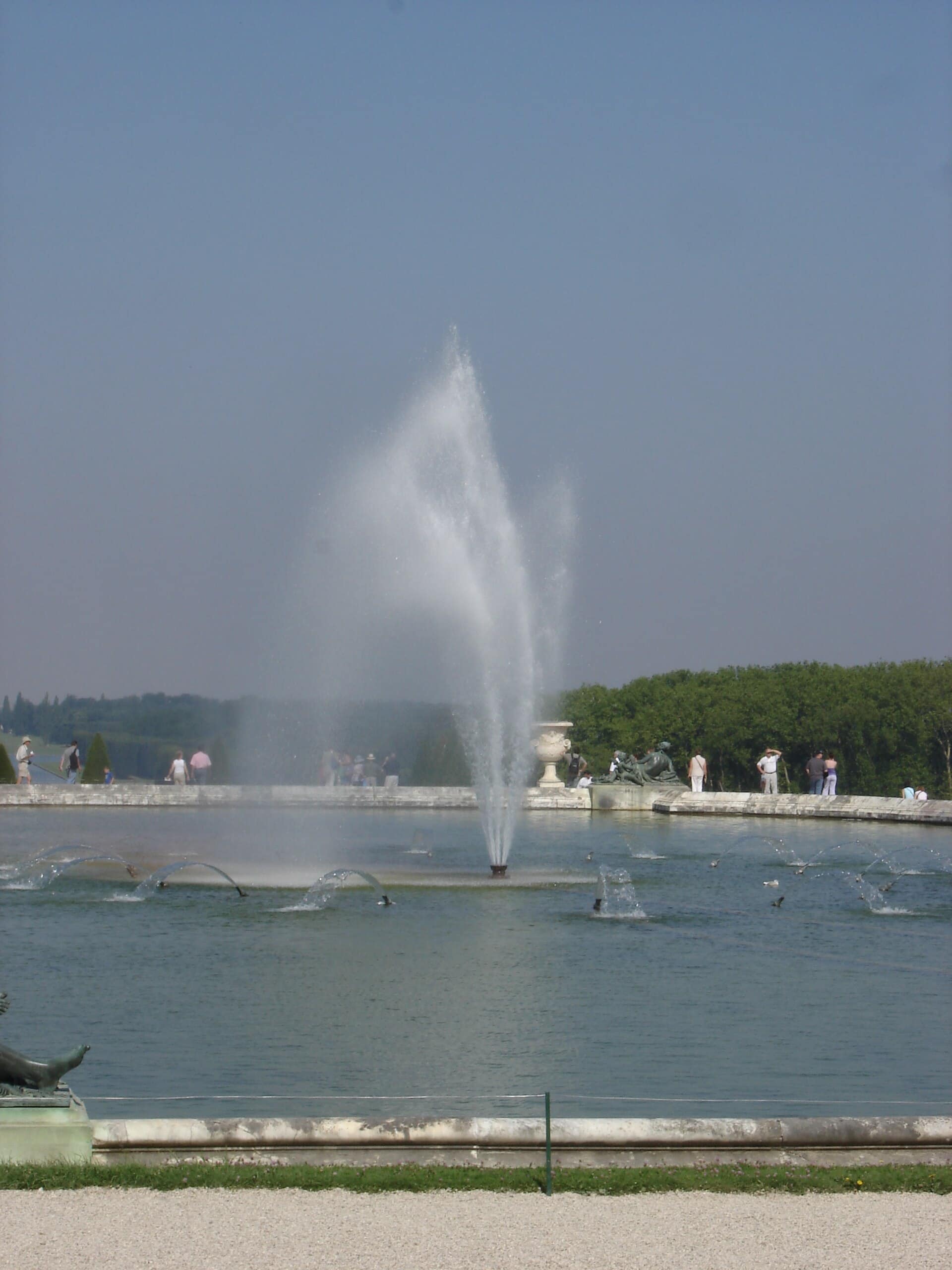 Un des deux bassins du parterre d’Eau de Versailles : grand bassin de pierre animé de jets d’eau, statue de bronze d’un fleuve couché sur la margelle, promeneurs alentour.