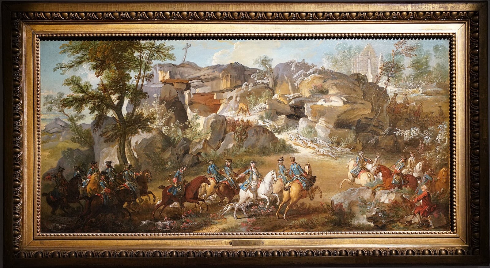 Tableau ancien encadré représentant une chasse royale à courre : cavaliers en livrée bleue et rouge rassemblés au premier plan, meute de chiens lancée entre des rochers, cerf fuyant sous une croix de pierre et les ruines d’une chapelle