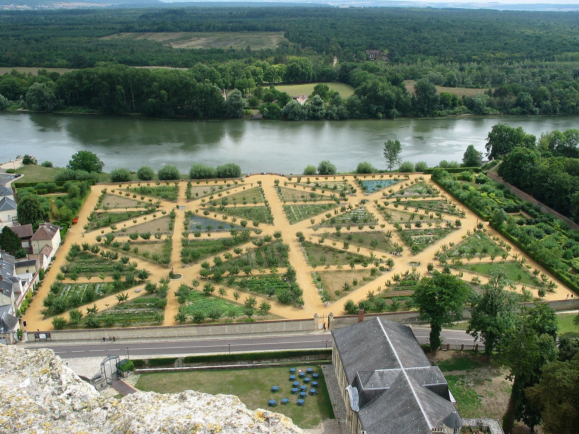 Vue aérienne du potager clos du château de La Roche-Guyon, au bord de la Seine : vaste enclos de murs divisé en compartiments triangulaires rayonnants, allées de sable ocre, arbres fruitiers alignés et bassins d’arrosage.