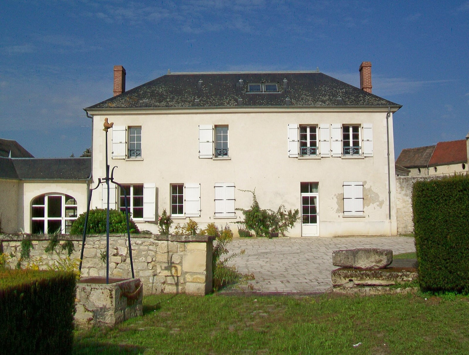 Ancien presbytère devenu mairie : bâtiment enduit à deux niveaux, façade régulière à volets blancs, toit à croupes d’ardoise et cheminées de brique, précédé d’un puits à potence de fer forgé et d’un muret de pierre dans un jardin.