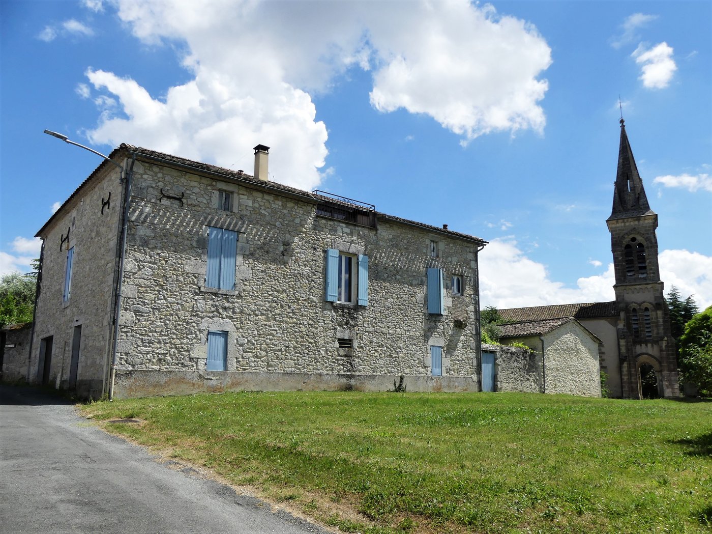 Presbytère en pierre calcaire à volets bleus et jardin, avec le clocher de l’église voisine visible à droite, à Lalandusse.
