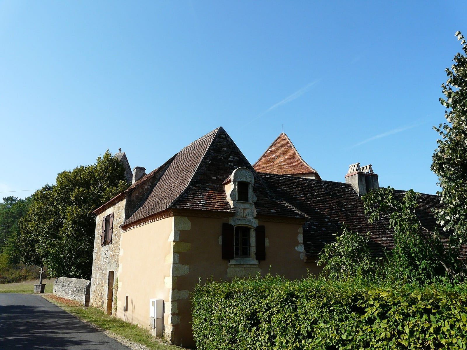 Ancien presbytère rural en pierre enduite aux angles de pierre de taille, toits pentus à croupes couverts de tuiles, fenêtre à encadrement mouluré et fronton à coquille, bordé d’une haie et d’une croix de chemin le long d’une route de village.