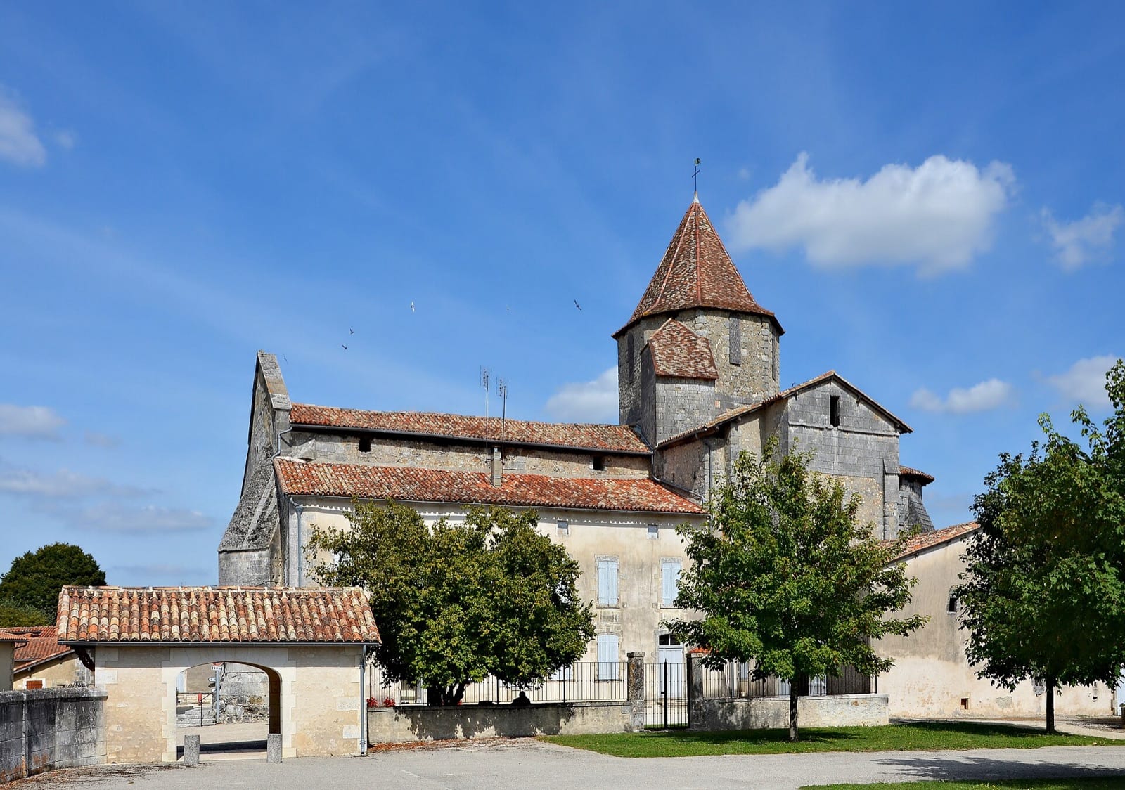 Église romane à clocher octogonal de pierre coiffé d’une flèche conique de tuiles, jouxtée par un presbytère à deux niveaux enduit aux volets bleus, séparés du parvis par un muret à grille, au cœur d’un bourg.