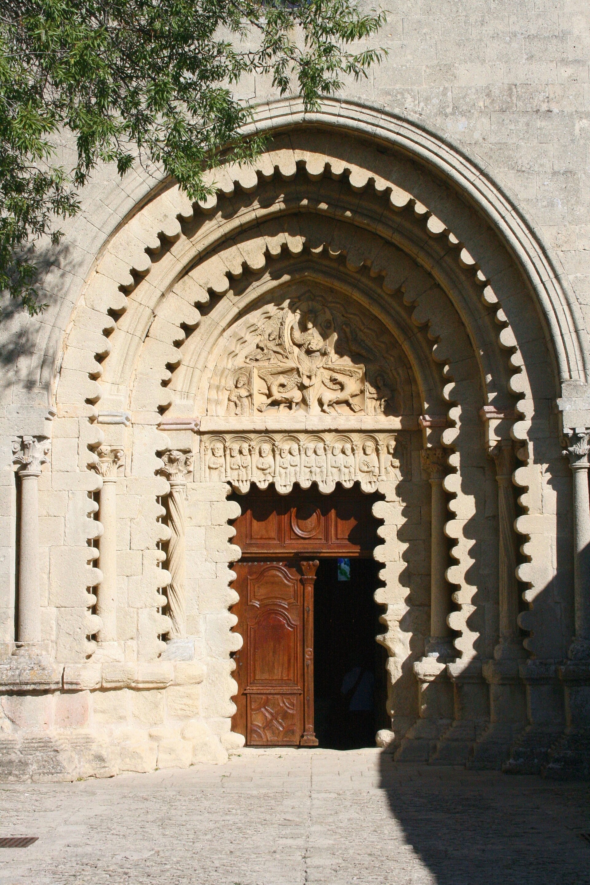 Portail roman d’une église : arc brisé aux voussures ornées d’un motif festonné en dents de scie, tympan sculpté d’un Christ entouré d’anges et de bêtes ailées, linteau à frise de petits personnages, colonnettes à chapiteaux, porte de bois, feuillage en haut à gauche