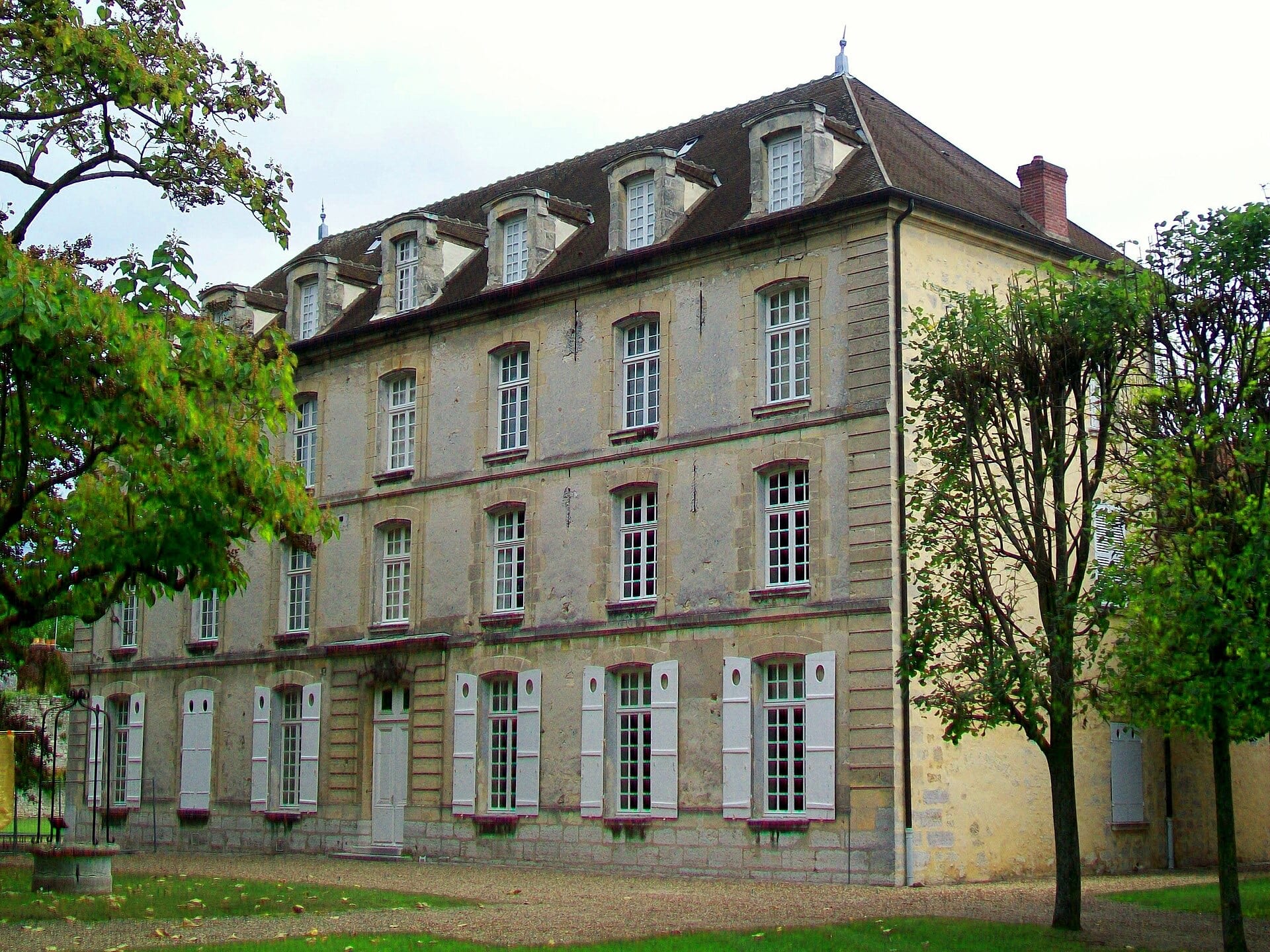 Grand logis classique de trois niveaux vu de biais : chaînes d’angle en pierre de taille, trois rangs de fenêtres à petits carreaux, lucarnes à fronton sur un haut toit de tuiles en pavillon, façades encadrées d’arbres