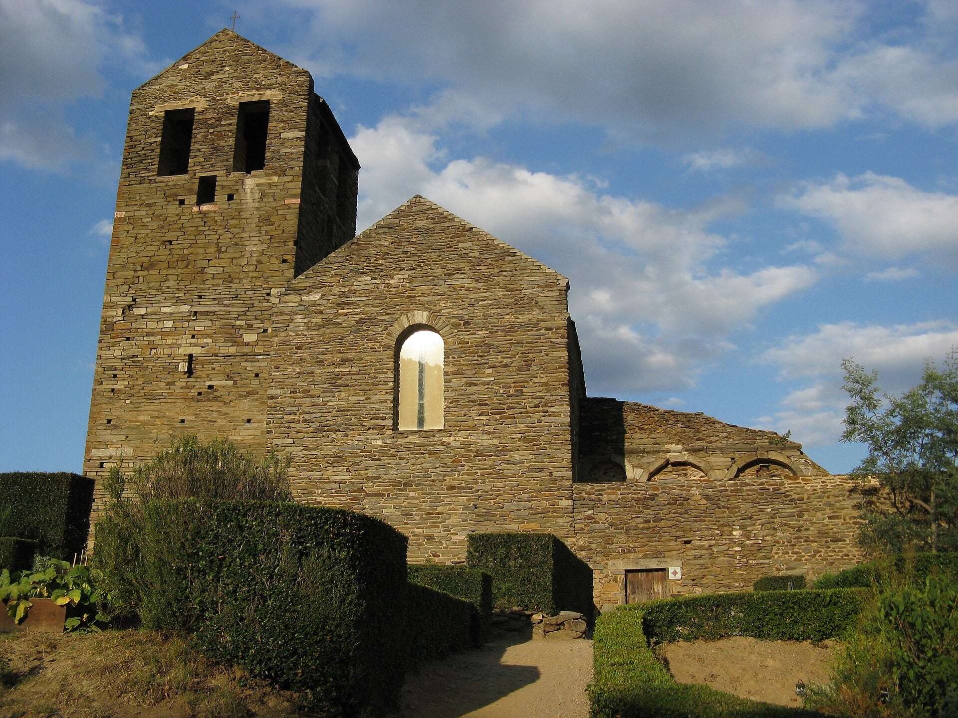 Petite église romane bâtie en pierres de schiste sombre : clocher-tour carré à baies géminées à gauche, nef à pignon percée d’une haute fenêtre étroite, murs bas et haies taillées au premier plan, ciel bleu
