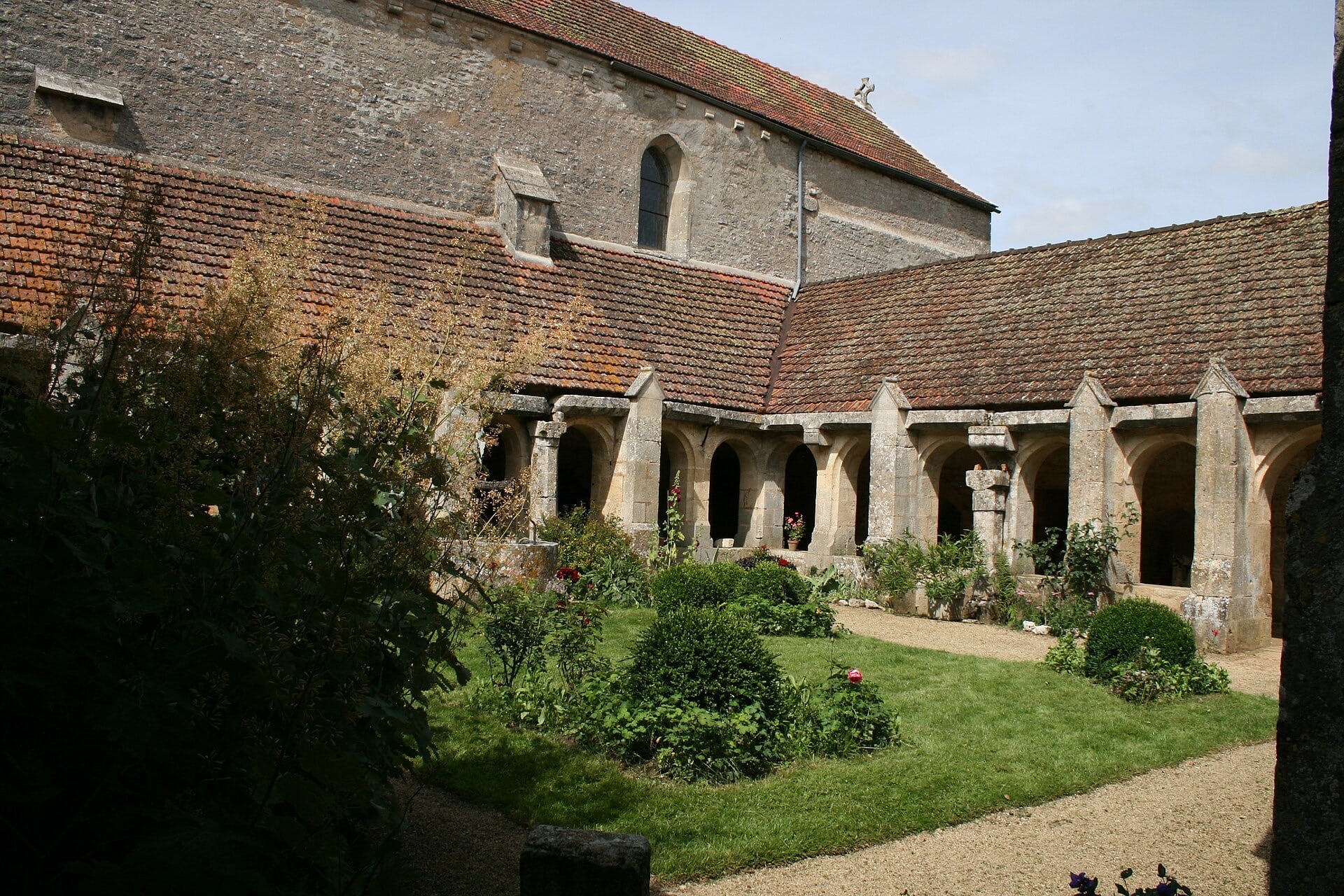 Galerie de cloître aux arcades en plein cintre appareillées de pierre, sous un toit de tuiles en appentis, autour d’un jardin planté ; au premier plan un puits de pierre circulaire surmonté d’un treuil de fer et de sa chaîne