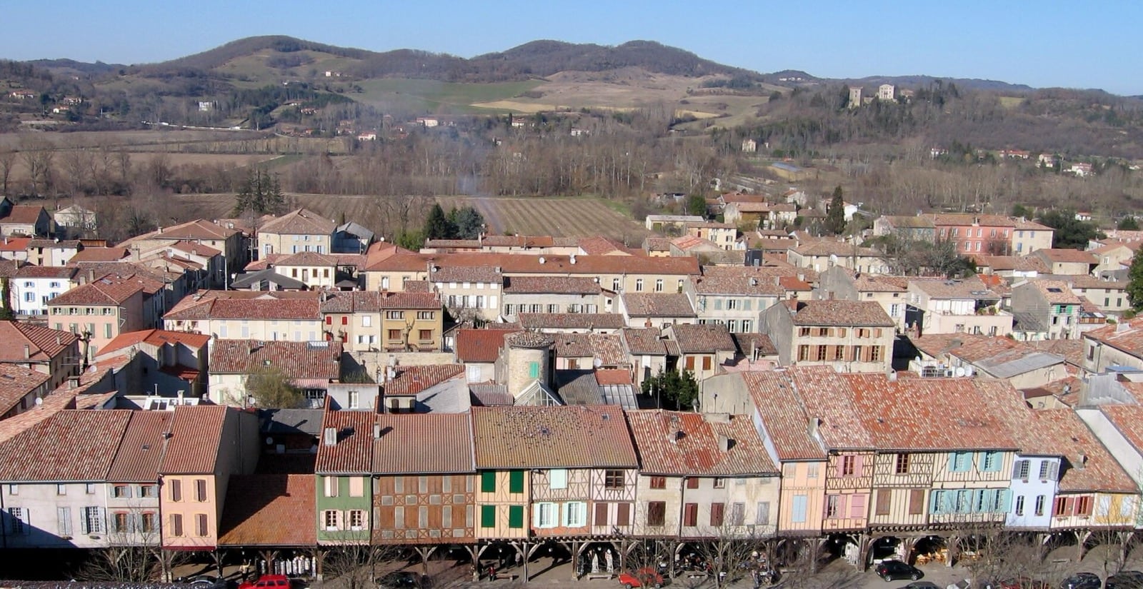 Mirepoix vue d'en haut : maisons à pans de bois colorés bordant la place, couverts de bois au rez-de-chaussée, plaine et collines au fond