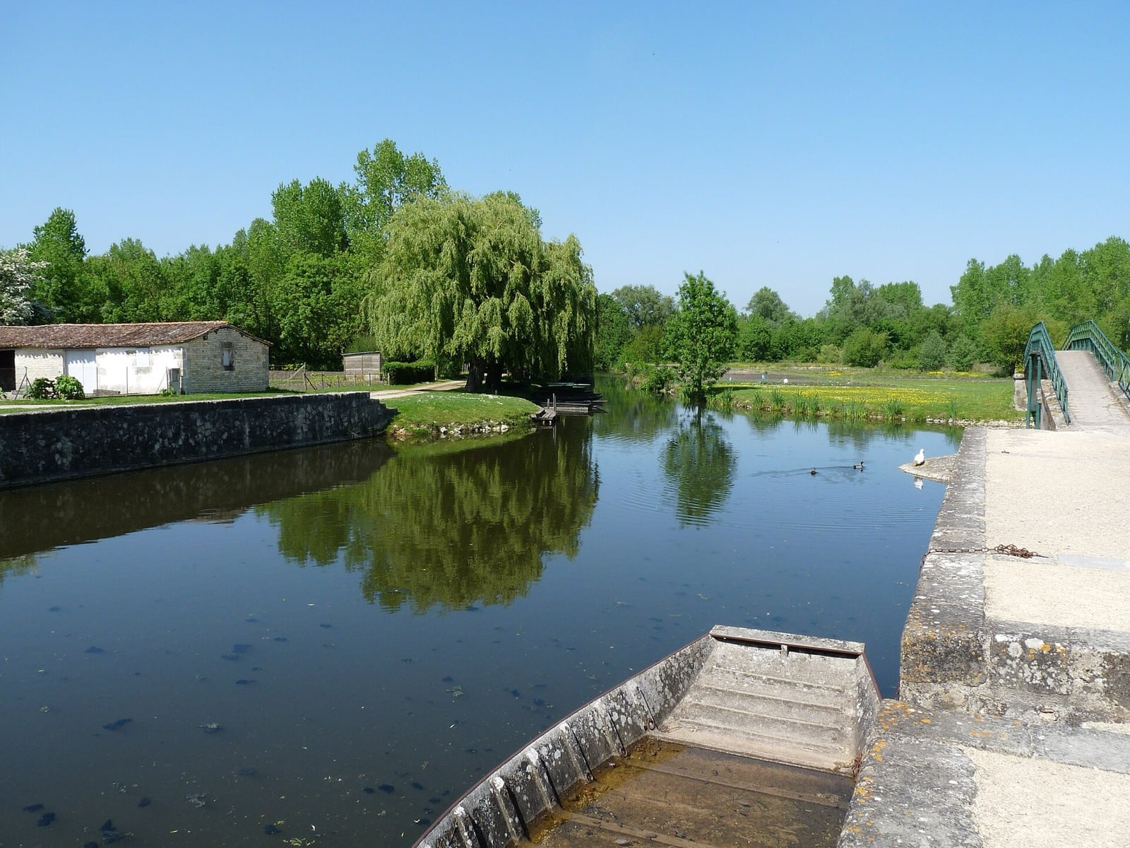 Port fluvial du Vanneau-Irleau : conche bordée d'un quai maçonné, barque à fond plat amarrée, passerelle métallique et saule pleureur