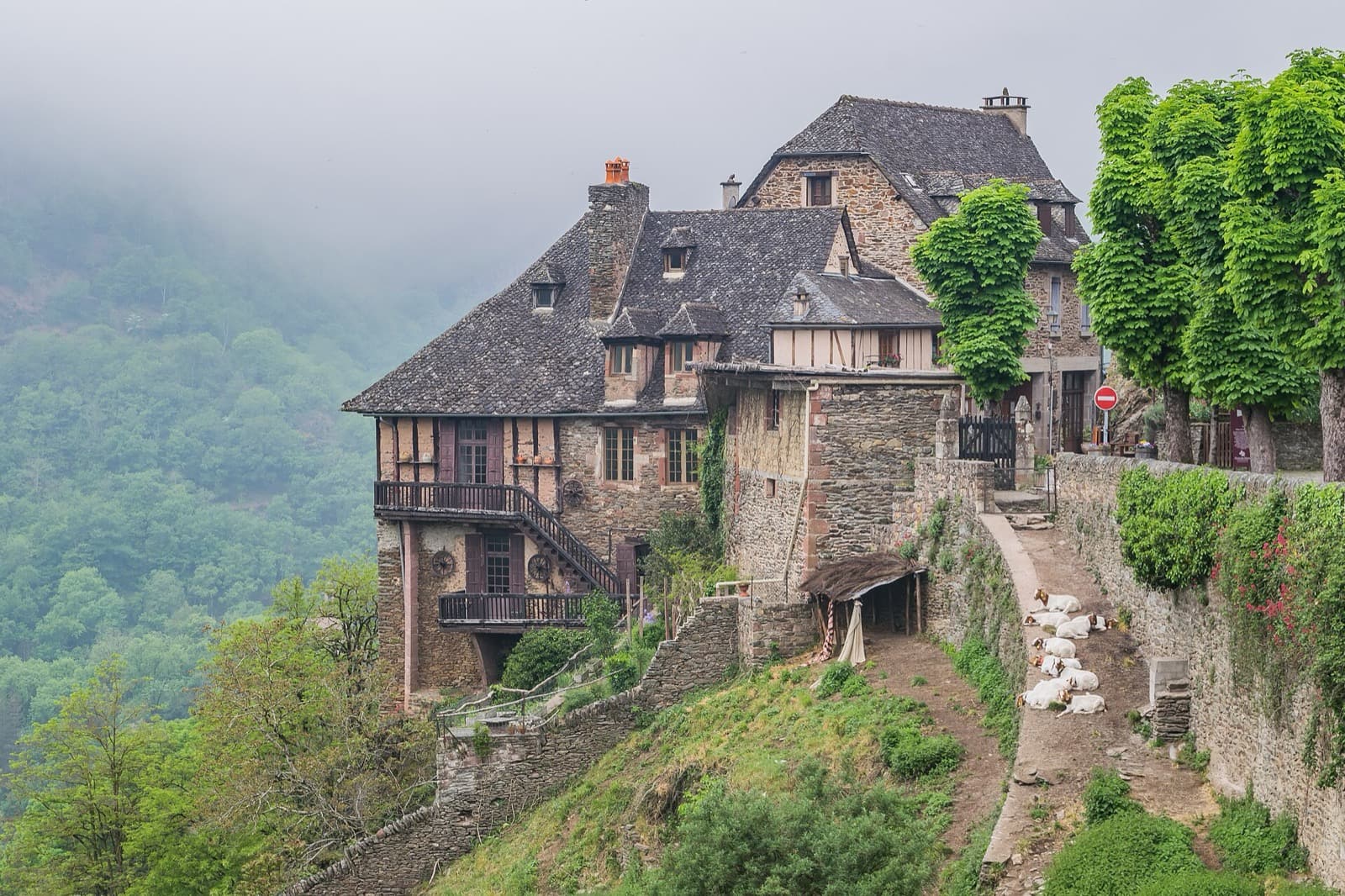 Maison de Conques en schiste, sous des toits de lauze de schiste à forte pente, dominant une vallée boisée dans la brume