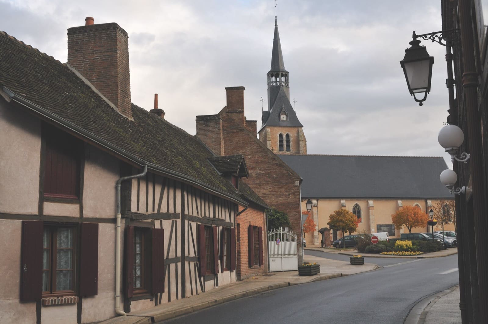 Rue de Chaumont-sur-Tharonne : maison à pan de bois, hautes souches de cheminée en brique, clocher à flèche d'ardoise au fond