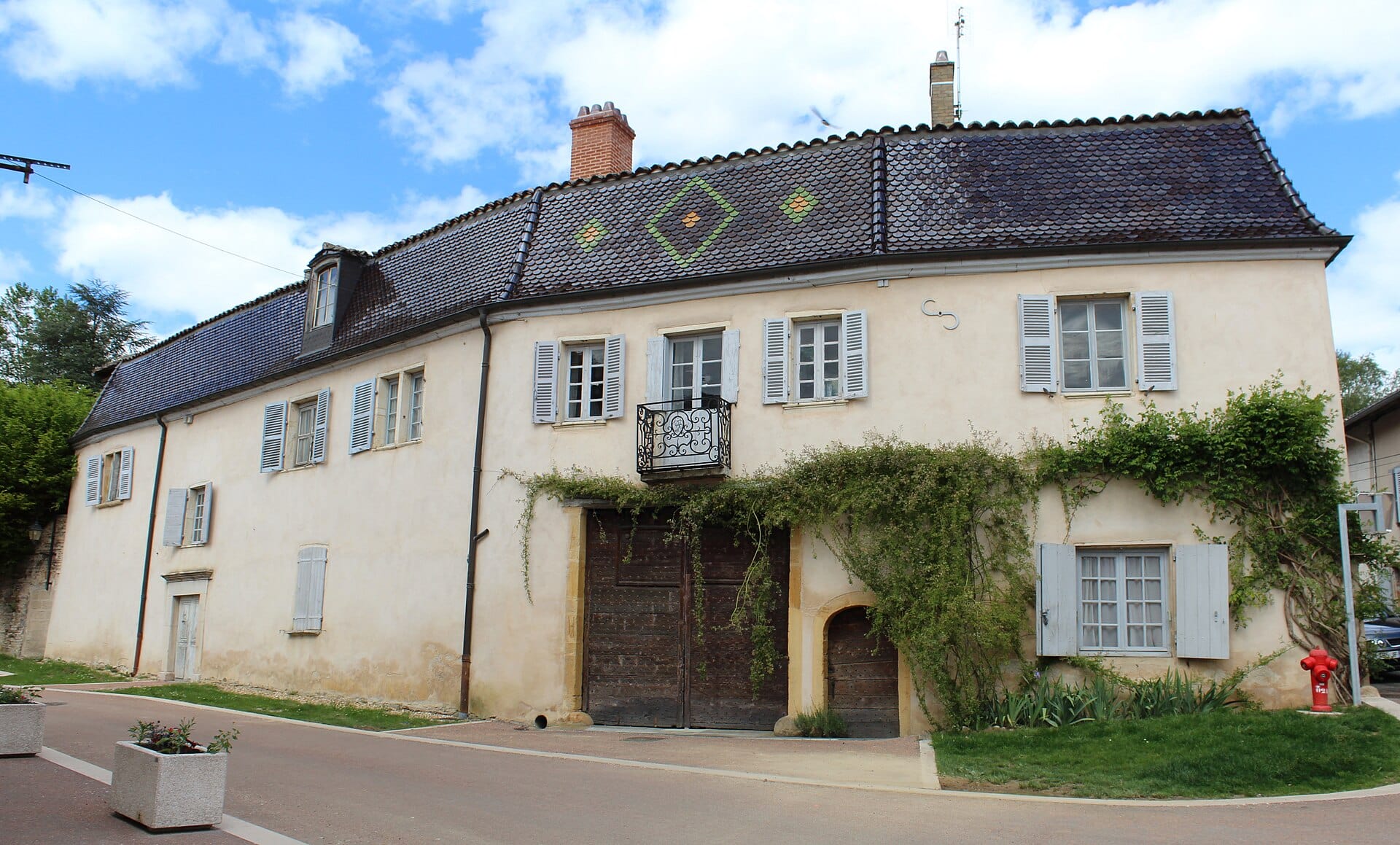 Ancien relais de poste de Guéreins : bâtiment enduit percé d’une large porte charretière à double vantail de bois, toiture à tuiles écaille aux losanges colorés, volets gris et vigne courant sur la façade.