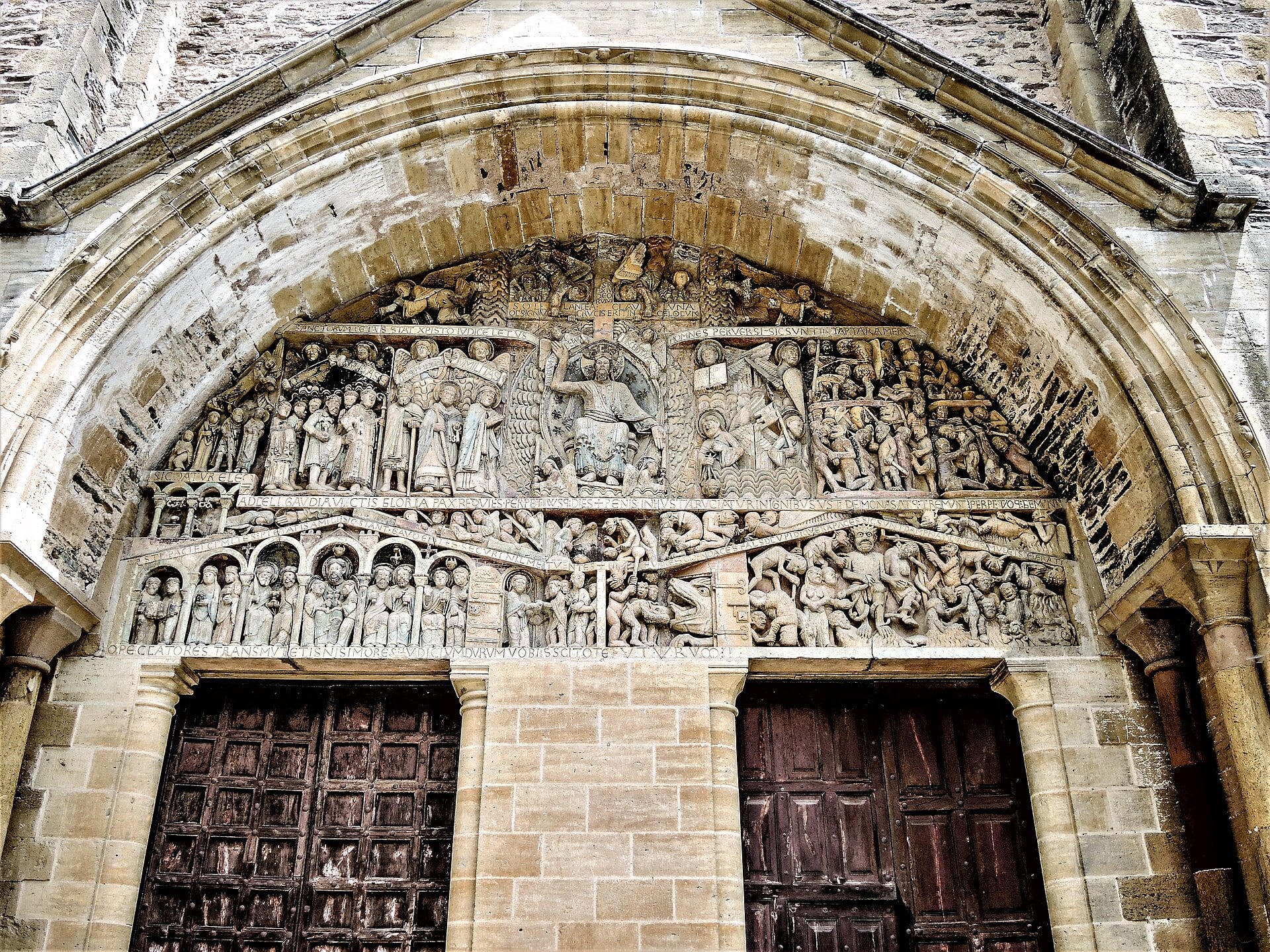 Tympan roman du Jugement dernier de l’abbatiale Sainte-Foy de Conques : le Christ en gloire dans une mandorle au centre, la main droite levée vers les élus rangés à gauche, la gauche abaissée vers les damnés livrés aux démons à droite, bandeaux couverts d’inscriptions latines
