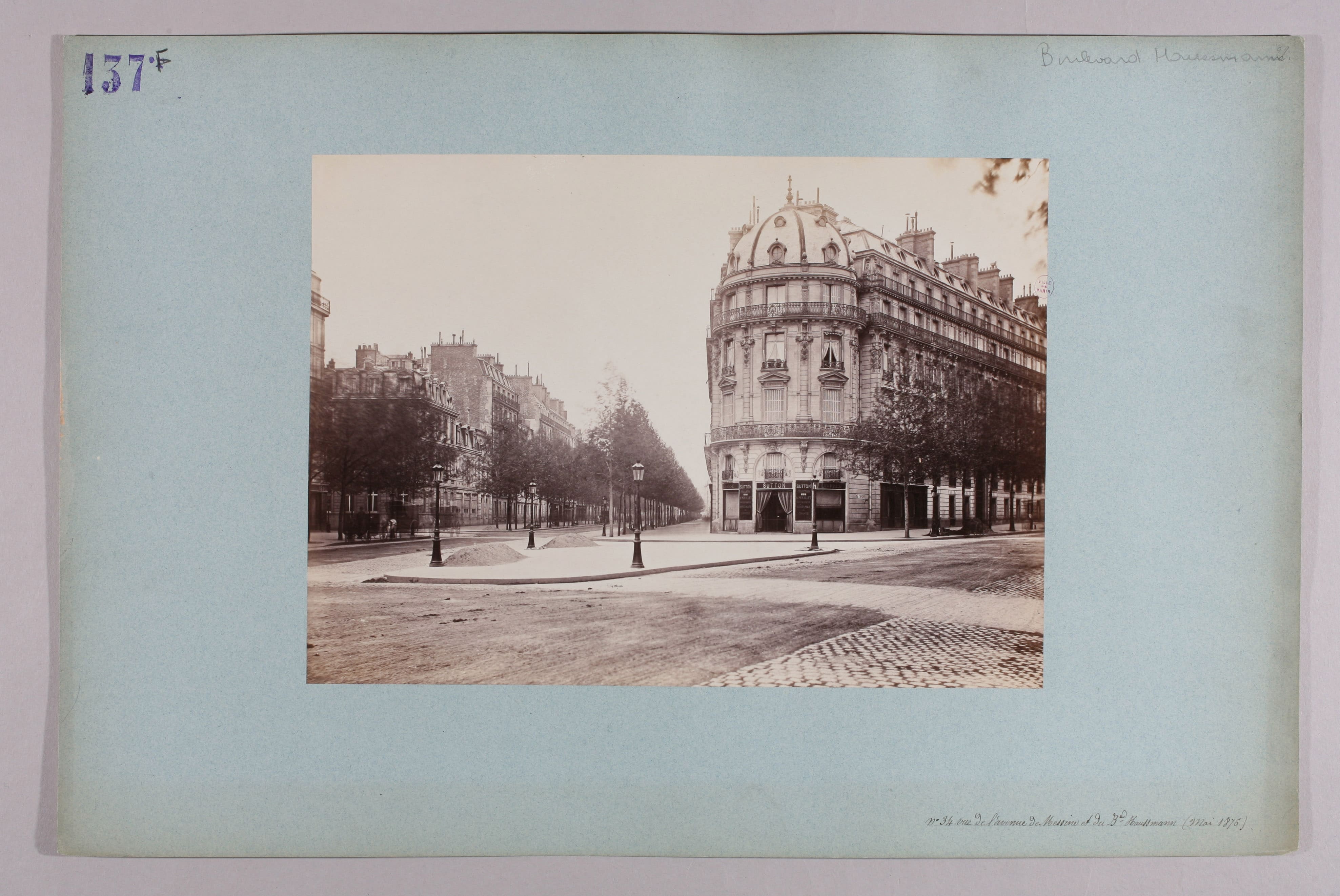 Photographie ancienne du boulevard Haussmann en 1876 : un immeuble d’angle à rotonde coiffée d’un dôme, une longue perspective bordée d’arbres et de réverbères, la chaussée en partie pavée