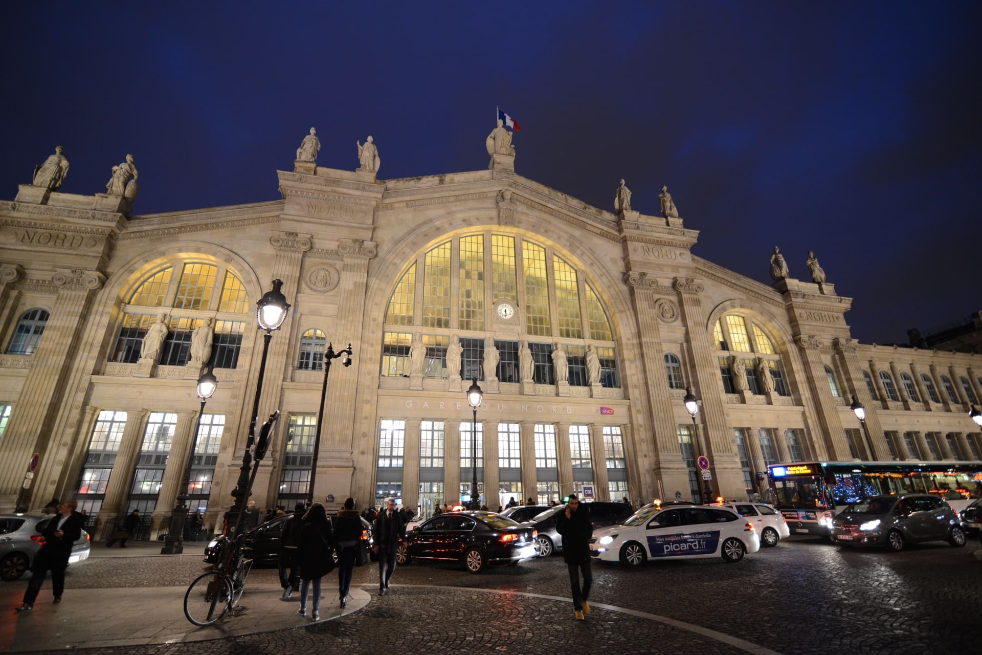La façade de la gare du Nord illuminée à la tombée de la nuit : grand arc vitré central, colonnes engagées, et une rangée de statues des villes desservies au sommet