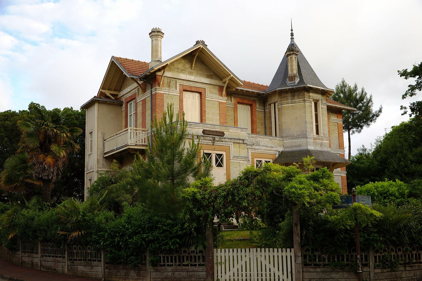 Villa de pierre claire et de brique rouge à composition asymétrique, tourelle d’angle coiffée d’un toit d’ardoise conique surmonté d’un épi, balcon de bois portant la plaque « Fragonard », sous des pins
