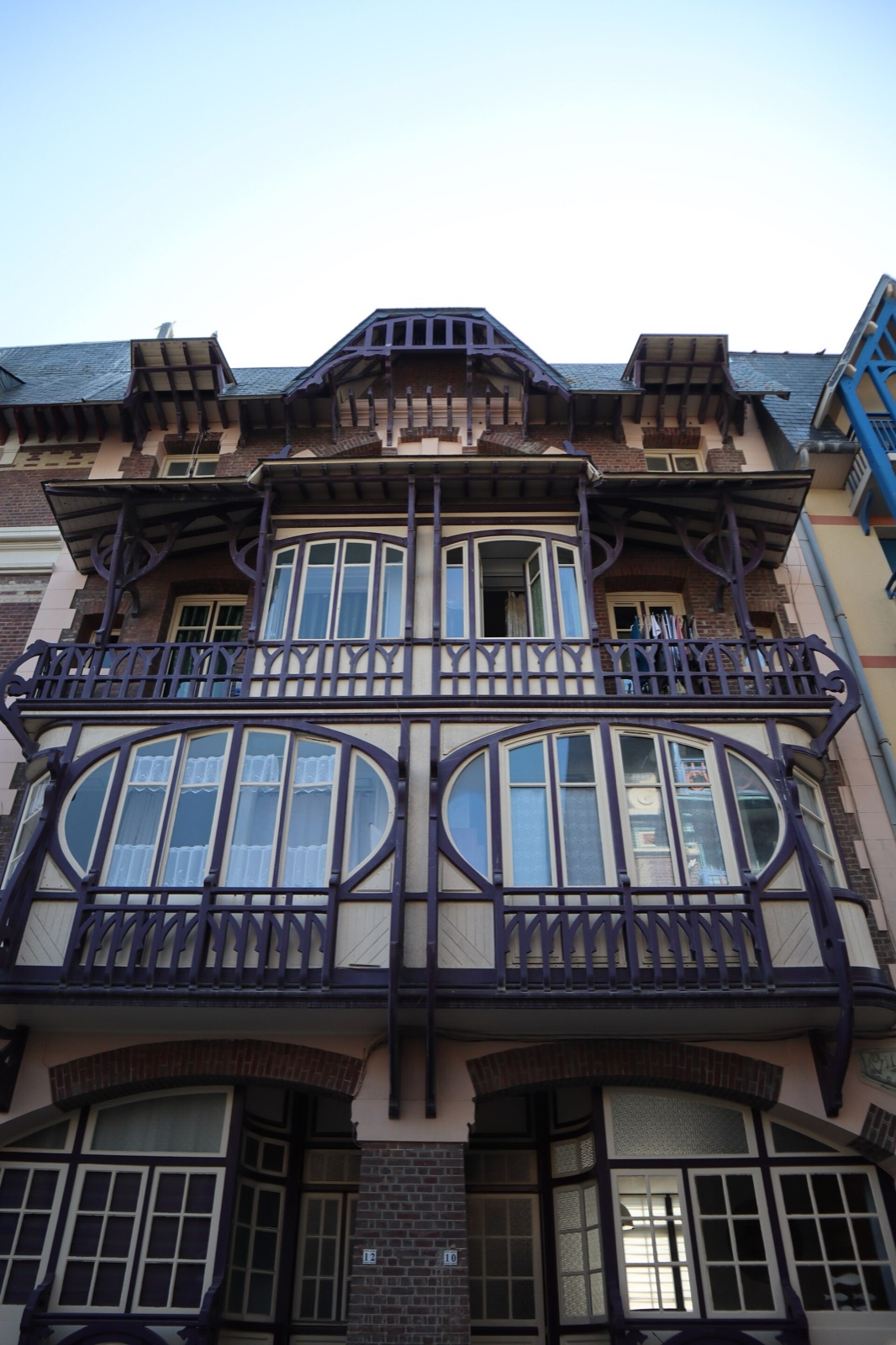 Façade de brique vue en contre-plongée : bow-windows vitrés à panneaux ovales superposés, loggias et balcons à balustres de bois peints en violet aux motifs courbes, aisseliers sculptés sous les débords de toit