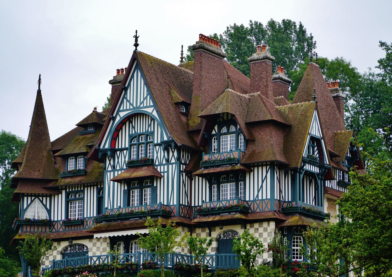 Grande villa à pans de bois peints en bleu-gris sur enduit blanc, toitures de tuiles très pentues à pignons découpés, tourelles à toits coniques, hautes souches de cheminées de brique, balcons de bois fleuris