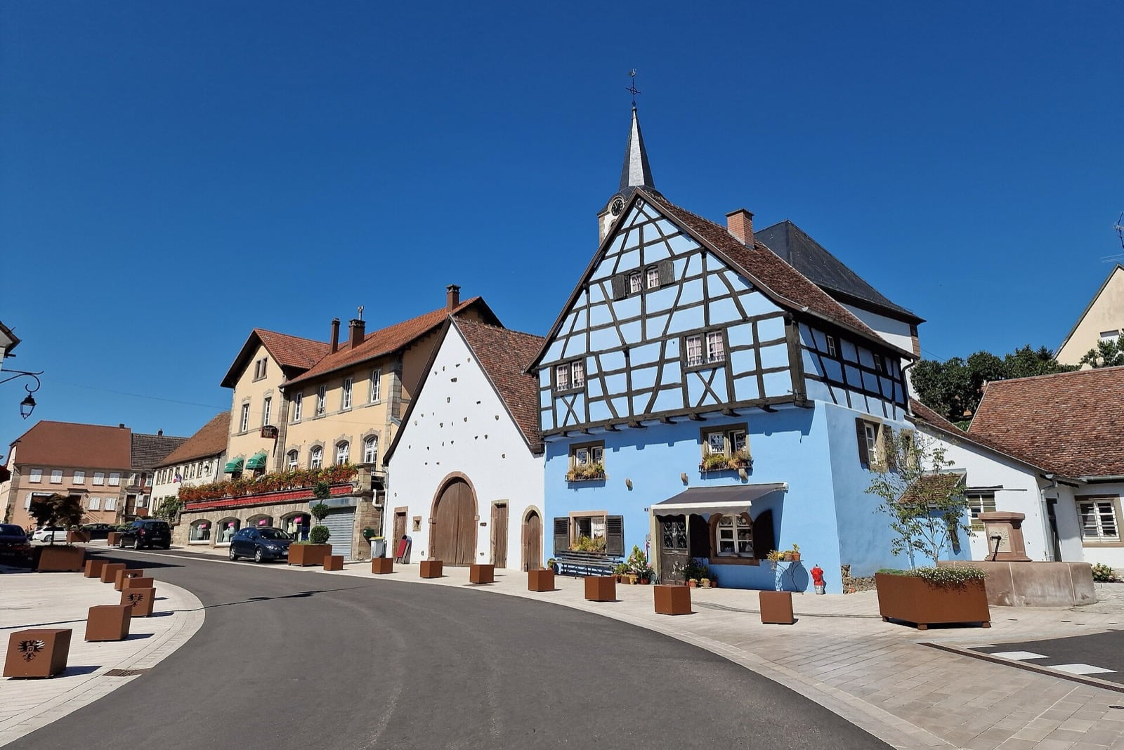 Cœur de village de Drulingen : maison à colombage bleue à tourelle d’horloge, alignée en front continu sur la rue