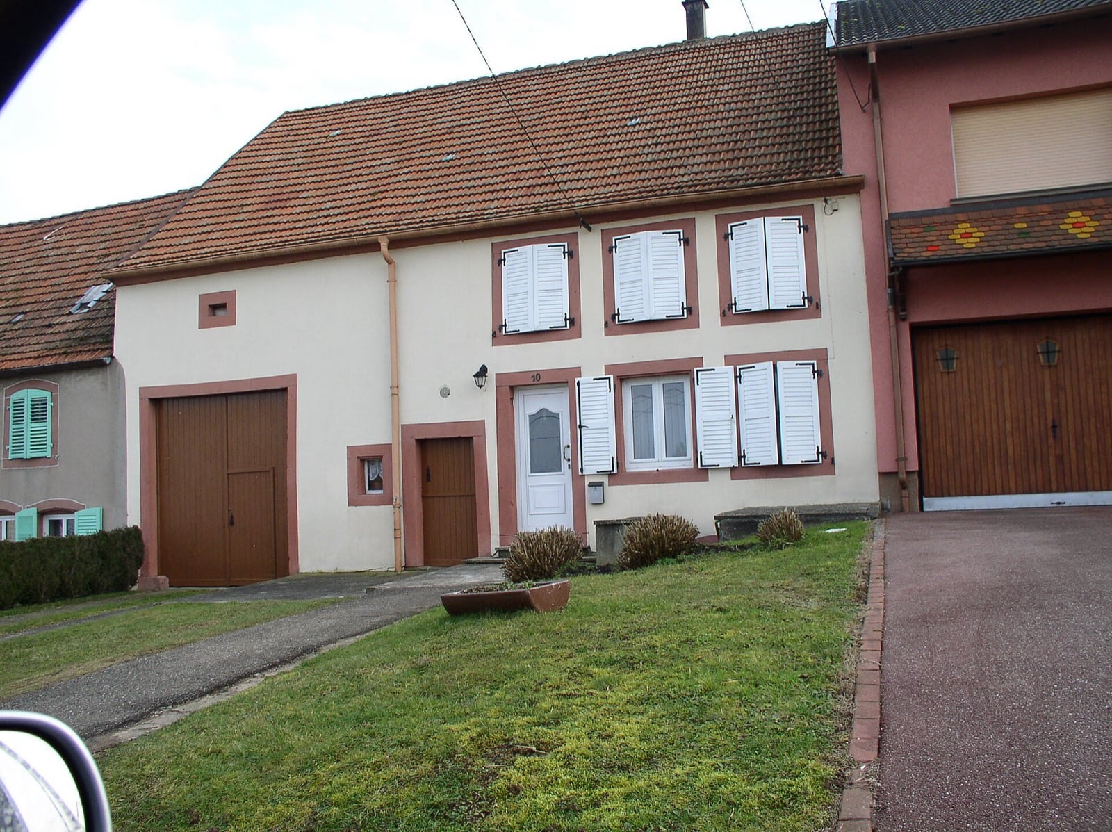 Maison lorraine à large porte de grange et porte piétonne, précédée d’une bande enherbée séparant la façade de la chaussée