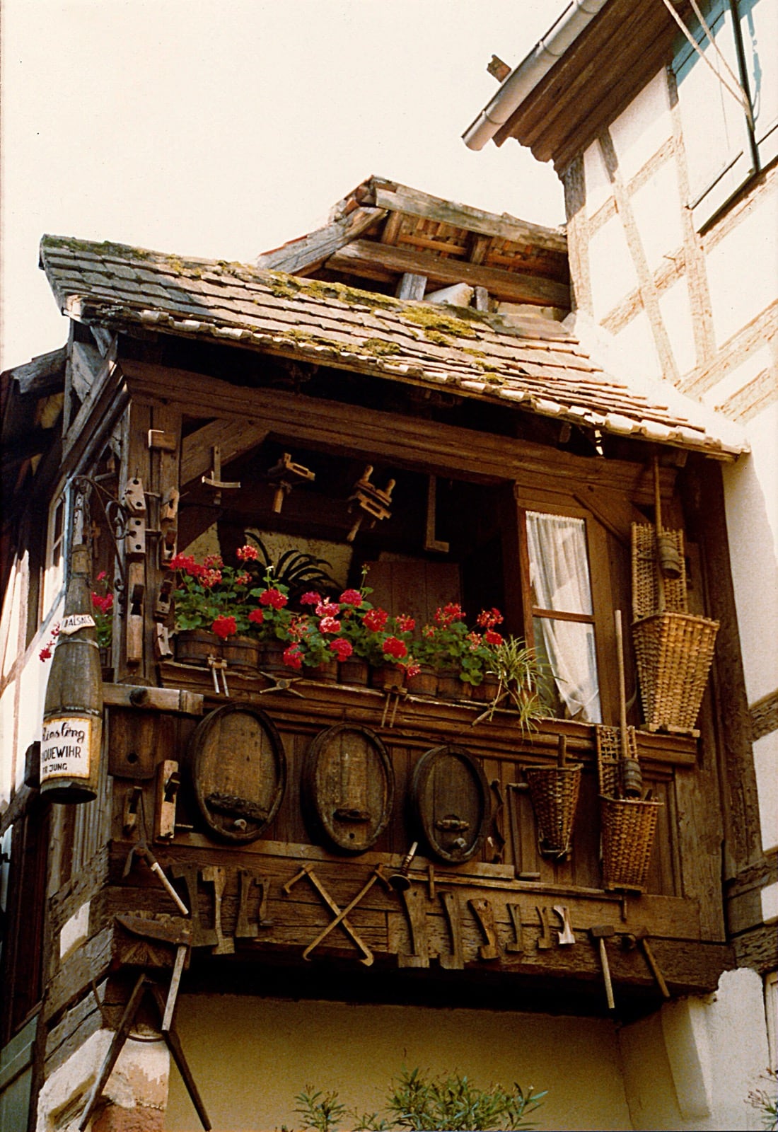 Balcon de bois d’une maison de vigneron à Riquewihr : tonneaux, paniers de vendange et outils de tonnellerie suspendus sous l’avant-toit