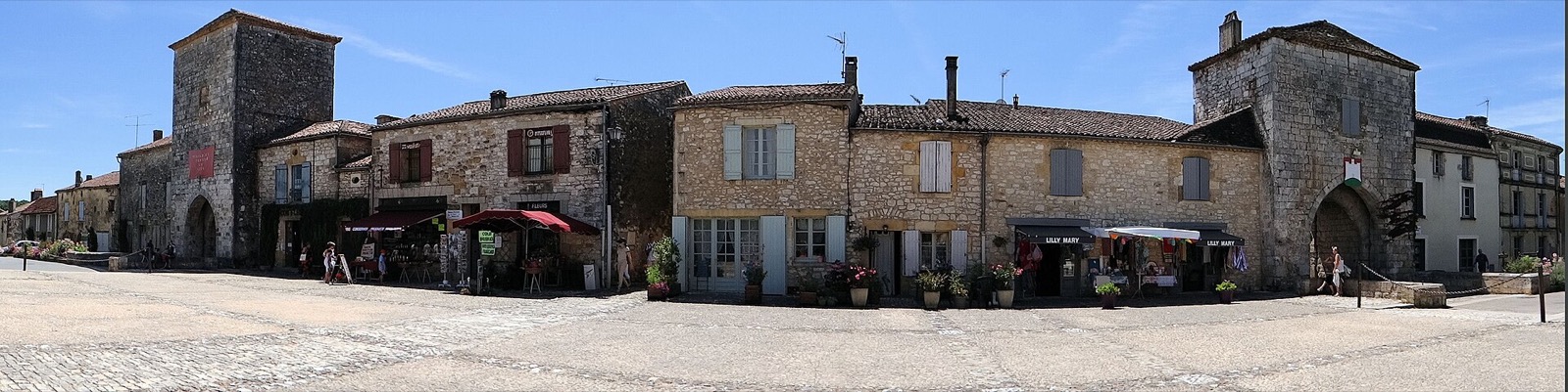 Monpazier au niveau de la rue : deux tours-portes de pierre encadrant une rue bordée d’arcades commerçantes