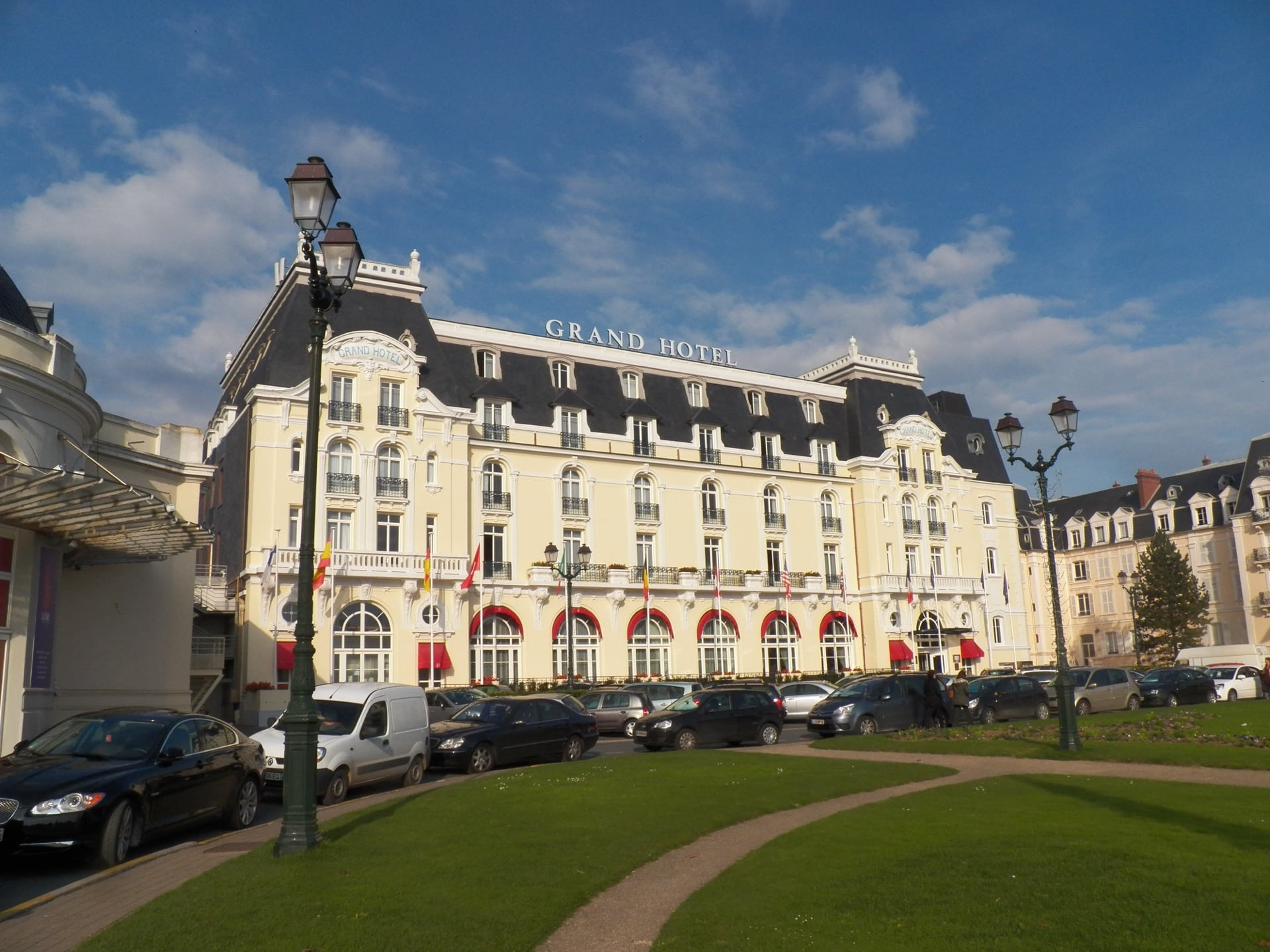 Le Grand Hôtel de Cabourg : longue façade classique claire à toiture Mansart d’ardoise, portant l’enseigne « Grand Hotel », arcades à stores rouges au rez-de-chaussée
