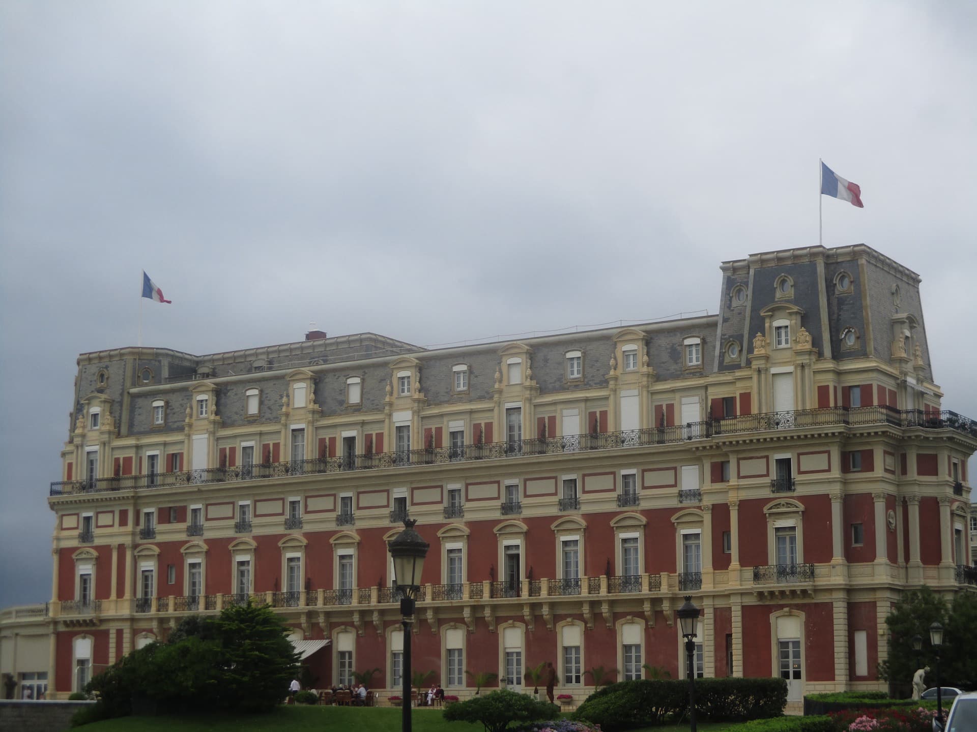 L’Hôtel du Palais de Biarritz : vaste palace de briques rouges et pierre blanche à toiture d’ardoise, pavillon d’angle surmonté d’un drapeau tricolore, sous un ciel gris